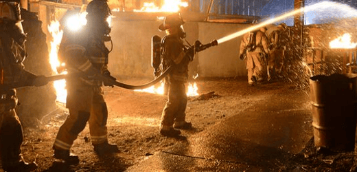 three fire fighters surrounded by a flaming building support a fire hose while the front person aims the water